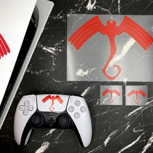 May include: A white video game controller with a red and black graphic of a stylized bird with wings spread. The controller is on a black surface with two smaller stickers of the same bird design.