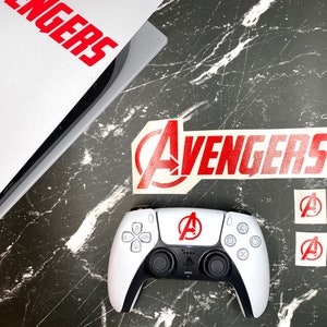 May include: A white PlayStation 5 DualSense controller with red Avengers logos on the controller and on stickers. The controller is on a black and white marble surface.