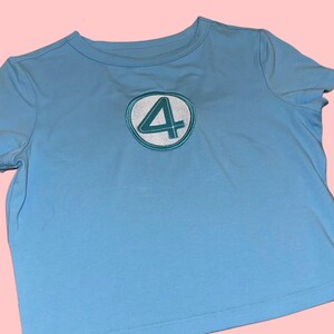 Peut inclure: T-shirt bleu clair avec un &eacute;cusson circulaire portant le chiffre "4" en blanc, bord&eacute; de turquoise. Le t-shirt a des manches courtes et un col rond. Le v&ecirc;tement est en tissu doux.