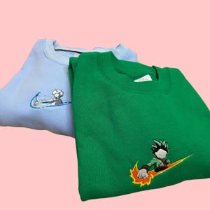 May include: Two crewneck sweatshirts, one light blue and one green, are displayed. Each sweatshirt features an embroidered design resembling a swoosh with a cartoon character. The light blue sweatshirt has a character surfing, while the green one has a character with fiery accents.