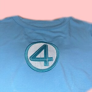 Peut inclure: T-shirt bleu clair avec un &eacute;cusson brod&eacute;. L'&eacute;cusson pr&eacute;sente le chiffre 4 en turquoise &agrave; l'int&eacute;rieur d'un cercle blanc, avec un contour turquoise. Le t-shirt a un col rond et est fait d'une mati&egrave;re douce.