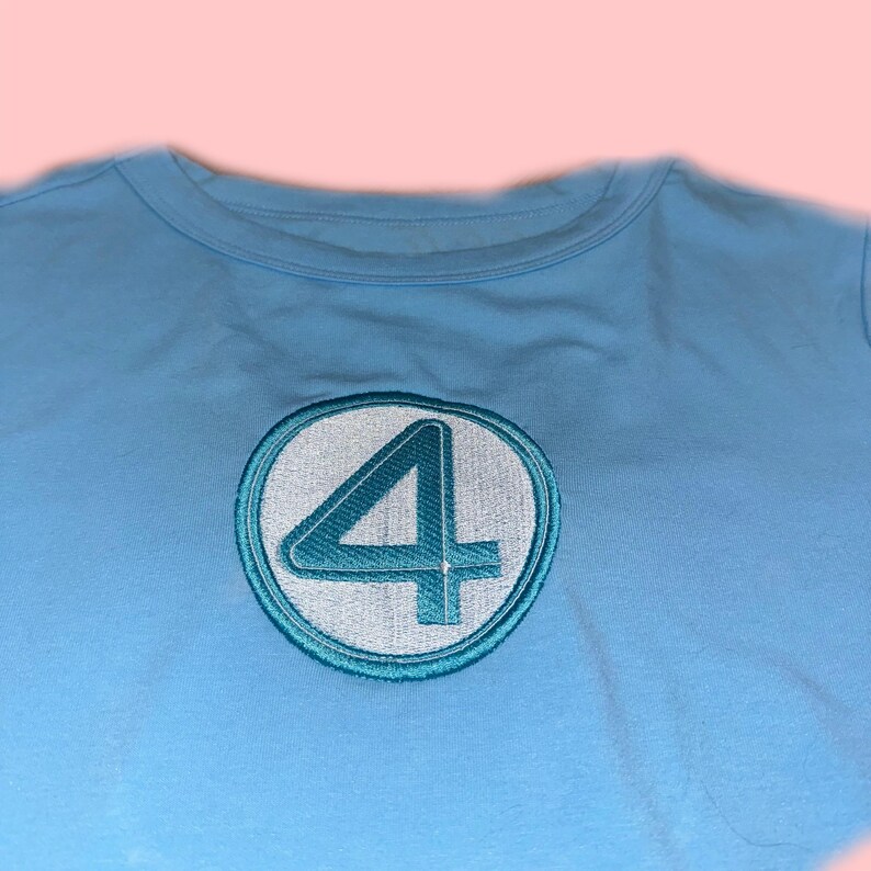Peut inclure: T-shirt bleu clair avec un &eacute;cusson brod&eacute;. L'&eacute;cusson pr&eacute;sente le chiffre 4 en turquoise &agrave; l'int&eacute;rieur d'un cercle blanc, avec un contour turquoise. Le t-shirt a un col rond et est fait d'une mati&egrave;re douce.