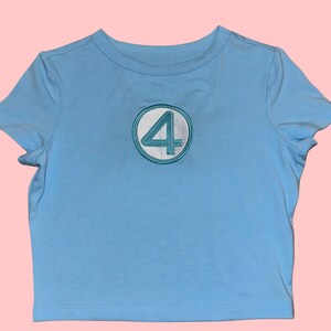 Peut inclure: T-shirt bleu clair &agrave; col rond et manches courtes. Le t-shirt pr&eacute;sente un embl&egrave;me circulaire avec une bordure blanche et le chiffre 4 turquoise au centre. Le t-shirt est en mati&egrave;re douce et convient aux tenues d&eacute;contract&eacute;es.