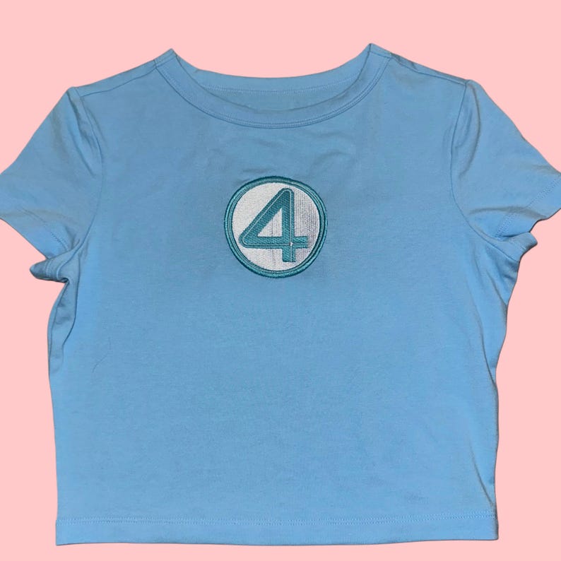 Peut inclure: T-shirt bleu clair &agrave; col rond et manches courtes. Le t-shirt pr&eacute;sente un embl&egrave;me circulaire avec une bordure blanche et le chiffre 4 turquoise au centre. Le t-shirt est en mati&egrave;re douce et convient aux tenues d&eacute;contract&eacute;es.