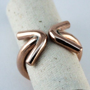 Può includere: Un anello in rame con un design unico che presenta due forme curve intrecciate. L'anello è lucido e ha una lucentezza metallica calda.