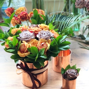 Peut inclure: Un bouquet de roses pêche, jaunes et violettes dans un vase en cuivre avec un ruban brun. Le vase est posé sur une table en bois avec deux vases en cuivre plus petits.