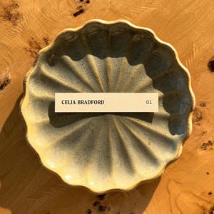 May include: A light gray ceramic dish with a scalloped edge. The dish is sitting on a wooden surface. A small, rectangular tag with the text "CELIA BRADFORD 01" is placed on the dish.