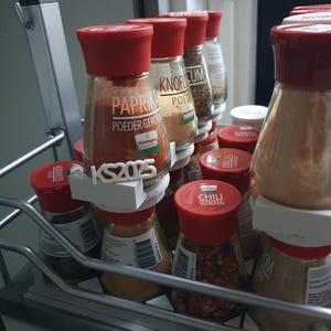 Peut inclure: Plusieurs pots d'épices avec des couvercles rouges, étiquetés avec des noms d'épices variés. Les pots sont disposés sur une étagère métallique. Les étiquettes incluent "Paprika", "Knoflook", et "Chili Vlokken".