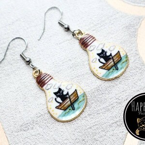 May include: A pair of silver earrings with a lightbulb charm. The lightbulb is white with a gold rim and features a black cat in a boat with clouds and a blue water scene.