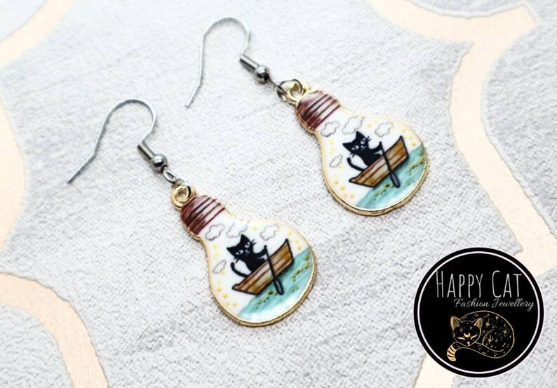 May include: A pair of silver earrings with a lightbulb charm. The lightbulb is white with a gold rim and features a black cat in a boat with clouds and a blue water scene.