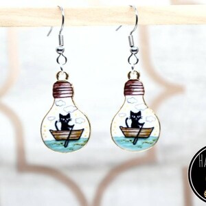 May include: A pair of gold lightbulb earrings with a black cat rowing a boat inside. The earrings have silver hooks.