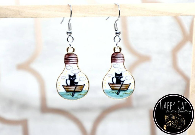 May include: A pair of gold lightbulb earrings with a black cat rowing a boat inside. The earrings have silver hooks.
