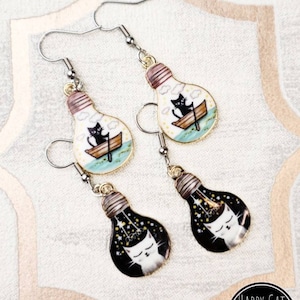 May include: A pair of silver dangle earrings featuring lightbulb charms. One charm has a black cat rowing a boat in a blue body of water with white clouds. The other charm has a white cat sleeping inside a black lightbulb with white stars.