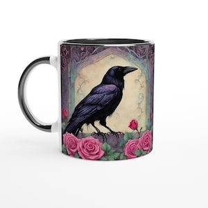 May include: A white ceramic mug with a black rim features a black raven perched on a stone ledge with pink roses in bloom. The background is a watercolour-style illustration of a stone archway with vines and a faded yellow background.