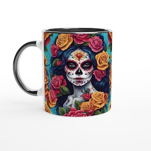 May include: White ceramic mug with a black rim and handle. The mug features a colorful illustration of a woman with a sugar skull makeup, surrounded by yellow, red, and pink roses on a turquoise background.