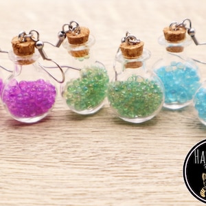 May include: Five pairs of glass bottle earrings with cork stoppers and silver hooks. The bottles are filled with iridescent beads in purple, green, blue, and light blue.