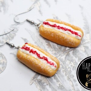 May include: Novelty earrings in the shape of miniature hot dogs. The earrings have a yellow bun, white cream, and red sauce, suspended from silver-coloured ear wires. The Happy Cat Fashion Jewellery logo is visible in the bottom right corner.