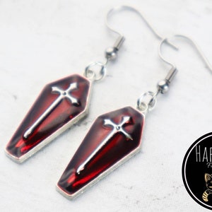 May include: A pair of silver earrings with red coffin-shaped charms and silver crosses. The earrings are on a white background.