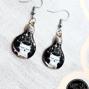 May include: A pair of silver earrings with a black and gold lightbulb design. The lightbulb features a white cat with closed eyes and gold stars inside. The earrings are labeled "Happy Cat Fashion Jewelry".