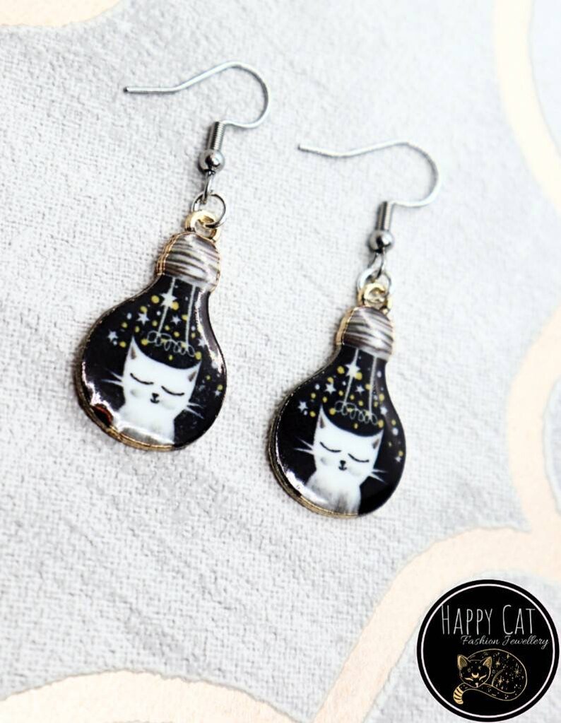 May include: A pair of silver earrings with a black and gold lightbulb design. The lightbulb features a white cat with closed eyes and gold stars inside. The earrings are labeled "Happy Cat Fashion Jewelry".