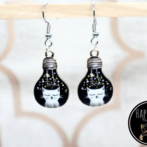 May include: A pair of silver dangle earrings with a black lightbulb charm featuring a white cat with closed eyes and gold stars. The lightbulb is surrounded by a black background with gold stars.