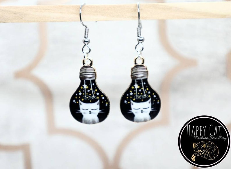 May include: A pair of silver dangle earrings with a black lightbulb charm featuring a white cat with closed eyes and gold stars. The lightbulb is surrounded by a black background with gold stars.