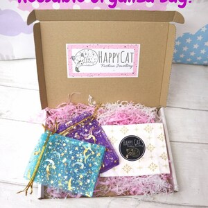 May include: A brown cardboard box with a white and pink tissue paper filler. The box contains two small organza bags with a moon and star pattern. The bags are labeled "HAPPYCAT Fashion Jewellery". The text "Plastic Free Packaging! Reusable Organza Bag!" is written in pink letters on a purple background at the top of the image. The text "*Packaging Colours May Vary*" is written in black letters on a white background at the bottom of the image.