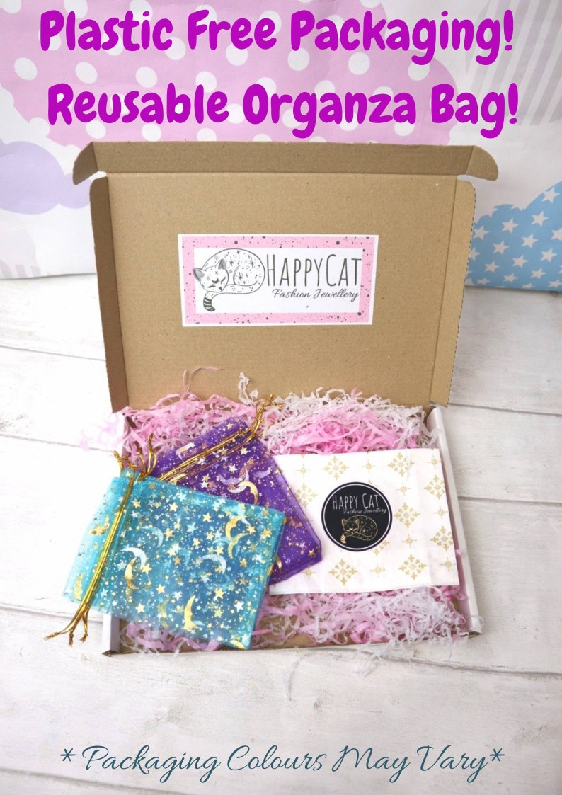 May include: A brown cardboard box with a white and pink tissue paper filler. The box contains two small organza bags with a moon and star pattern. The bags are labeled "HAPPYCAT Fashion Jewellery". The text "Plastic Free Packaging! Reusable Organza Bag!" is written in pink letters on a purple background at the top of the image. The text "*Packaging Colours May Vary*" is written in black letters on a white background at the bottom of the image.