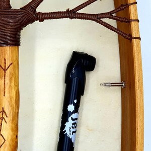 Puede incluir: Primer plano de un tambor de marco de madera con cordones de cuero marr&oacute;n. Una bomba de bicicleta negra con la palabra "Jet" y el texto "Zeital" est&aacute; en primer plano.