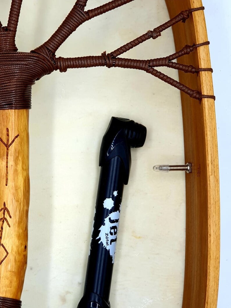 Puede incluir: Primer plano de un tambor de marco de madera con cordones de cuero marr&oacute;n. Una bomba de bicicleta negra con la palabra "Jet" y el texto "Zeital" est&aacute; en primer plano.