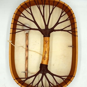 Puede incluir: Tambor de marco de madera de forma ovalada con una cabeza de cuero marr&oacute;n. El tambor tiene un dise&ntilde;o &uacute;nico en forma de &aacute;rbol con una cuerda marr&oacute;n tejida en el marco. Se incluye un palo de madera con una bola blanca en el extremo.