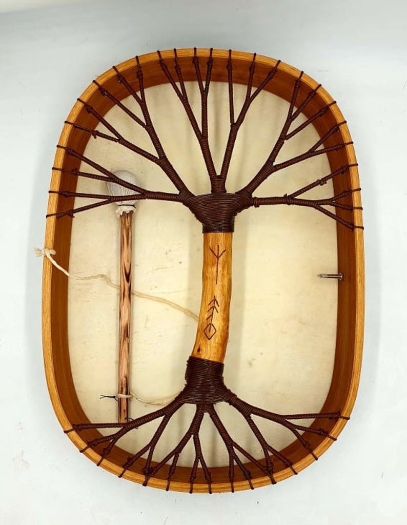Puede incluir: Tambor de marco de madera de forma ovalada con una cabeza de cuero marr&oacute;n. El tambor tiene un dise&ntilde;o &uacute;nico en forma de &aacute;rbol con una cuerda marr&oacute;n tejida en el marco. Se incluye un palo de madera con una bola blanca en el extremo.