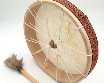 May include: A round wooden frame drum with a white leather head and a brown and orange patterned strap. The drum is accompanied by a wooden beater with a brown brush on one end.