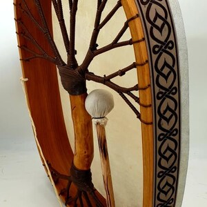Puede incluir: Un tambor de marco de madera marr&oacute;n con una cabeza de cuero blanco y una correa de cuero marr&oacute;n. El tambor tiene un dise&ntilde;o de &aacute;rbol de la vida en la parte delantera y un dise&ntilde;o de nudo celta en el lateral. El tambor est&aacute; siendo sostenido por un palo de madera con una cabeza de fieltro blanco.