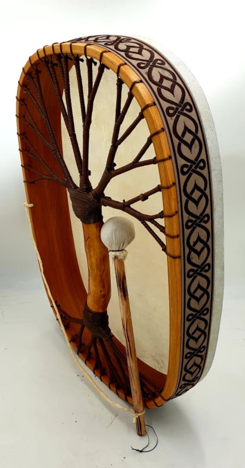 Puede incluir: Un tambor de marco de madera marr&oacute;n con una cabeza de cuero blanco y una correa de cuero marr&oacute;n. El tambor tiene un dise&ntilde;o de &aacute;rbol de la vida en la parte delantera y un dise&ntilde;o de nudo celta en el lateral. El tambor est&aacute; siendo sostenido por un palo de madera con una cabeza de fieltro blanco.