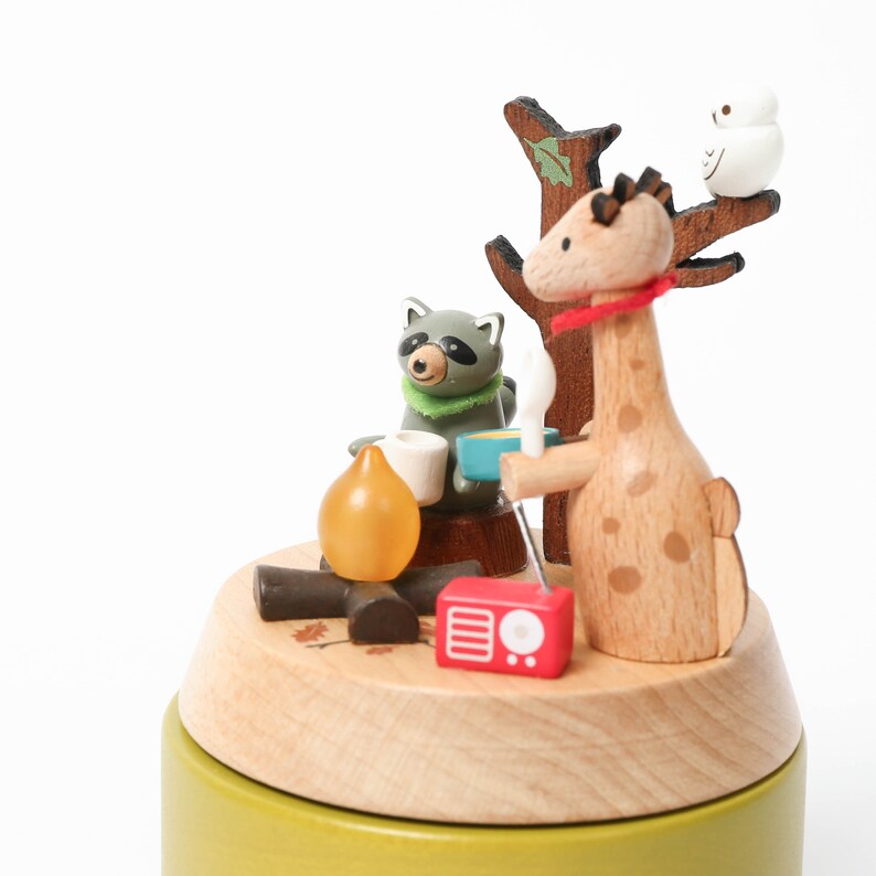 May include: A wooden music box with a giraffe, raccoon, and bird figurine. The giraffe is holding a cup and the raccoon is holding a cup and a marshmallow. The music box is playing a song.