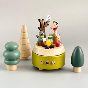 May include: A wooden music box with a green base and a scene of a giraffe, raccoon, and owl around a campfire. The music box has a wooden tree, a green tree, and a brown tree on the sides. The music box plays the song "FMA".