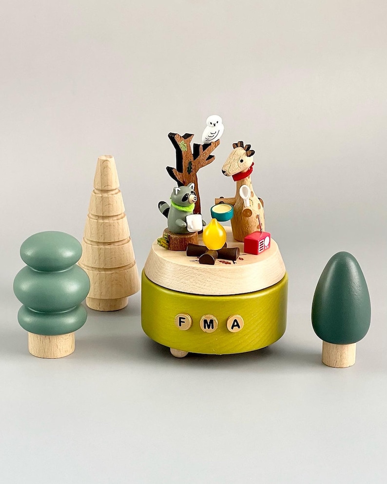 May include: A wooden music box with a green base and a scene of a giraffe, raccoon, and owl around a campfire. The music box has a wooden tree, a green tree, and a brown tree on the sides. The music box plays the song "FMA".