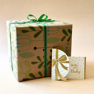 May include: A gift box wrapped in brown paper with a green and white patterned design. The box is tied with a green ribbon and bow. A gold card with a white and blue bow and the text "Hello! new Baby" is next to the box.