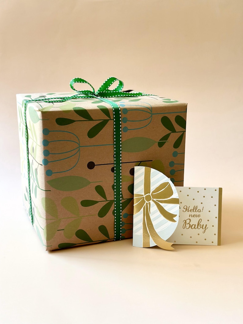 May include: A gift box wrapped in brown paper with a green and white patterned design. The box is tied with a green ribbon and bow. A gold card with a white and blue bow and the text "Hello! new Baby" is next to the box.