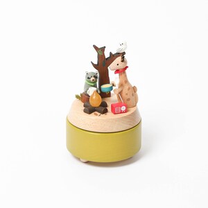 May include: A wooden music box with a giraffe, raccoon, and owl figurine on top. The music box is painted green and has a brown base. The figurines are all sitting around a campfire.