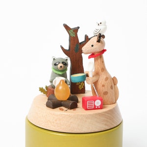 May include: A wooden music box with a giraffe, raccoon, and owl figurine. The giraffe is holding a spoon and a bowl. The raccoon is holding a cup. The owl is perched on the giraffe's head. The music box is decorated with a campfire, a tree, and a radio.