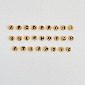 May include: A set of 26 gold-colored letter beads with black lettering. The beads are arranged in alphabetical order from A to Z.
