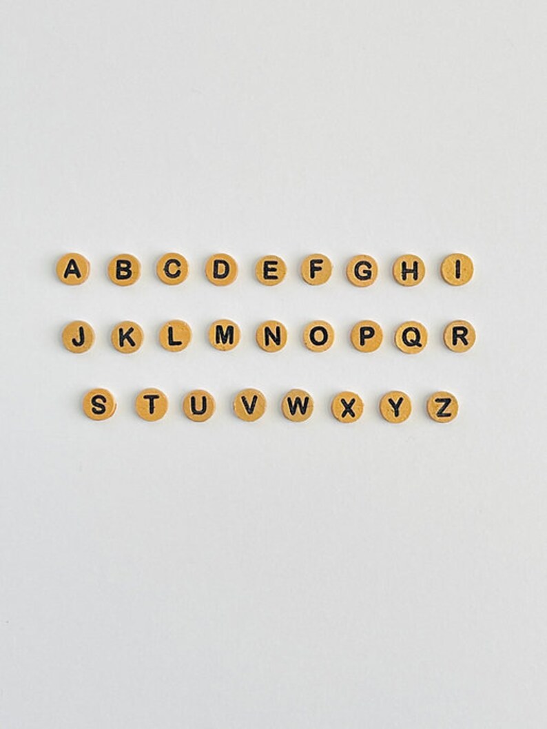 May include: A set of 26 gold-colored letter beads with black lettering. The beads are arranged in alphabetical order from A to Z.