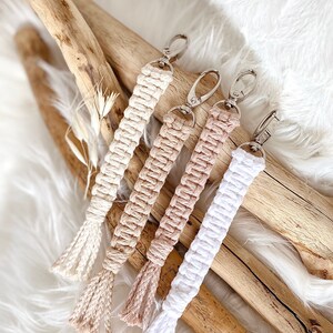 Könnte beinhalten: Vier Makramee-Schlüsselanhänger in Creme, Beige und Weiß, jeweils mit einem silbernen Verschluss und einer geknoteten Quaste. Die Schlüsselanhänger sind auf einer weißen, flauschigen Oberfläche mit Treibholz im Hintergrund angeordnet.