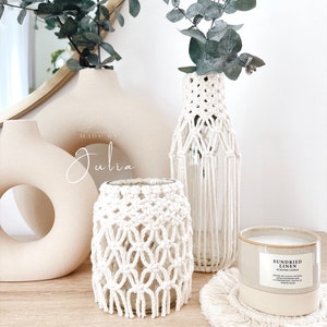 Op de afbeelding: Een verzameling woondecoratie-artikelen, waaronder een beige vaas, een met macramé omwikkelde glazen pot en een doorzichtige glazen fles met macramé details. Een geurkaars en een macramé onderzetter zijn ook aanwezig.