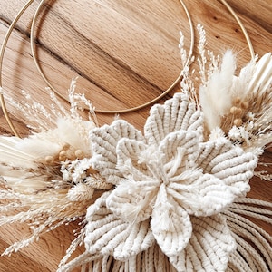 Op de afbeelding: Een macramé wandhanger met een grote, crèmekleurige bloem in het midden, omringd door gedroogde grassen en bloemen. Het ontwerp is geplaatst op een gouden metalen ring, met extra macramé franjes eronder. De natuurlijke texturen en neutrale tinten creëren een bohemien esthetiek.