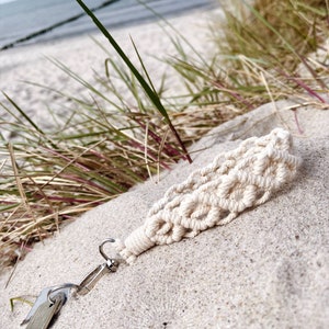 May include: A cream-colored macrame keychain with a silver-tone clasp and keys rests on sand. The keychain features a woven design and is near beach grass and the ocean. The text "made by Julia" is visible in the bottom right corner.