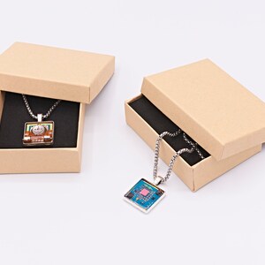 May include: Two brown cardboard boxes with lids open, each containing a silver chain necklace with a square pendant. The pendant in the left box has a colourful design, while the pendant in the right box has a blue and green design.