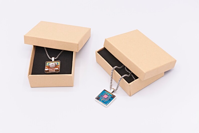 May include: Two brown cardboard boxes with lids open, each containing a silver chain necklace with a square pendant. The pendant in the left box has a colourful design, while the pendant in the right box has a blue and green design.